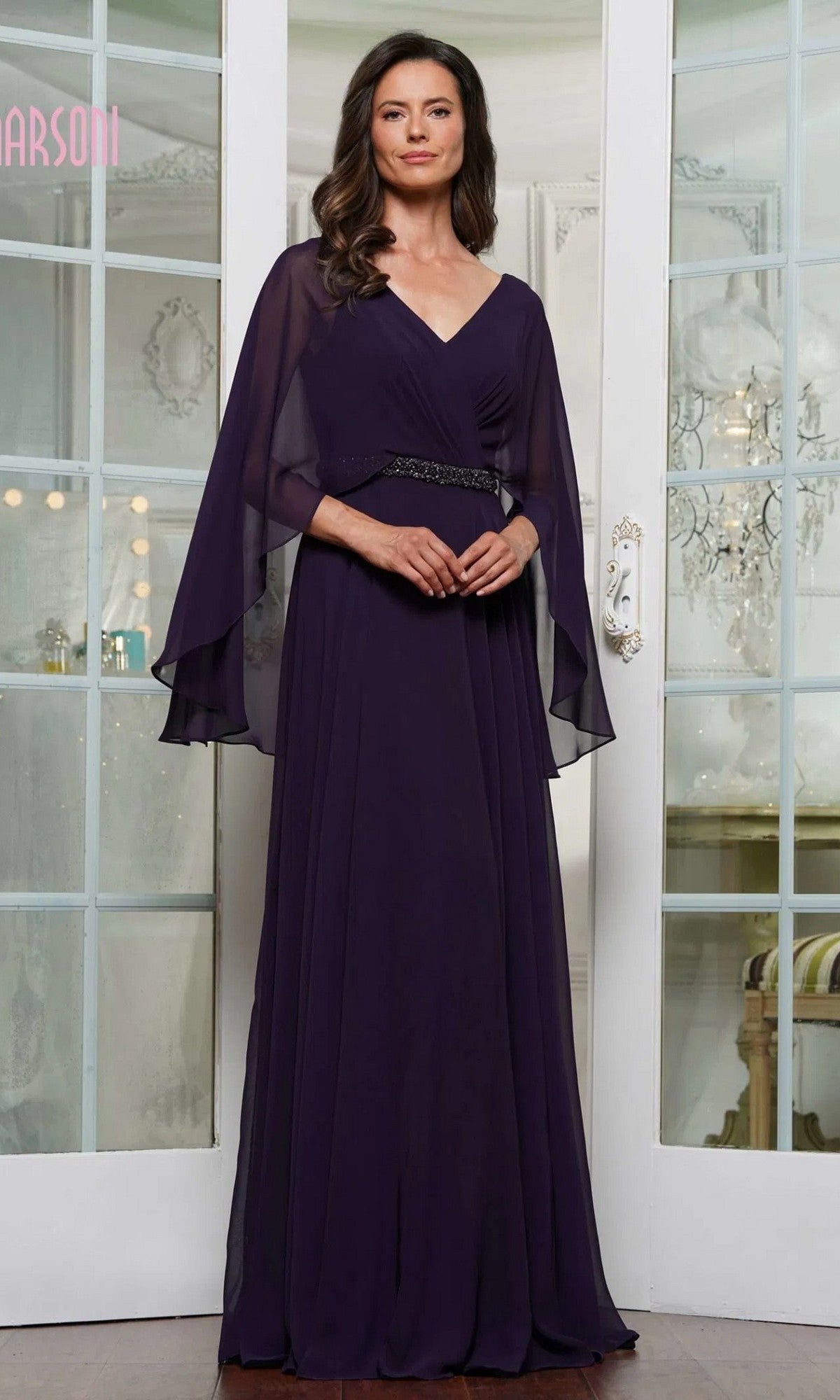 A woman poses indoors in front of glass-paneled doors, wearing the Marsoni MV1350 long formal dress in aubergine chiffon with a sheer capelet and an embellished waistbelt.
